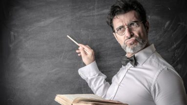 science teacher with a book at a blackboard thinking of answer to question